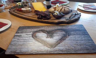 A laid table with a wooden board full of cheese, sausage, and bread. In the foreground is a placemat with a heart pattern.