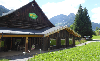 Ein rustikales Holzgebäude mit einem grünen Schild. Im Hintergrund sind hohe Berge und ein blauer Himmel zu sehen. | © iKuh Schöne Ideen | Daniela Neumann