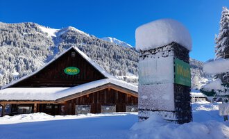 Ein gemütliches Holzhaus im Schnee mit einem klaren blauen Himmel im Hintergrund. Die Umgebung ist von verschneiten Bergen und Wäldern umgeben.