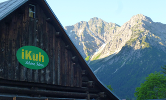 Ein traditionelles Holzhaus mit einem grünen Schild, das "iKuh" zeigt. Im Hintergrund sind majestätische Berge und ein klarer Himmel zu sehen. | © iKuh Schöne Ideen | Daniela Neumann