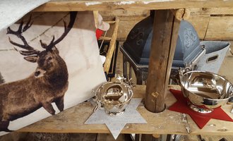 Eine rustikale Dekoration mit einem Kissen, das ein Bild eines Hirsches zeigt, und glänzenden Metallschalen. Die Schalen stehen auf roten und grauen Sternen auf einem Holztisch.