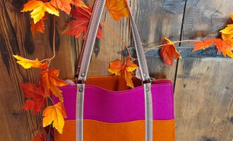 Eine stilvolle Tasche in Orange und Pink hängt vor einer rustikalen Holzoberfläche. Rote und orangefarbene Blätter umrahmen das Bild und verleihen eine herbstliche Atmosphäre. | © iKuh Schöne Ideen | Daniela Neumann