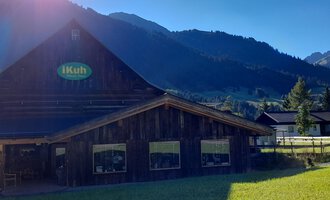 Ein Holzgebäude mit einem Schild "iKuh" in einer grünen Wiese. Im Hintergrund sind Berge und ein klarer blauer Himmel sichtbar. | © iKuh Schöne Ideen | Daniela Neumann