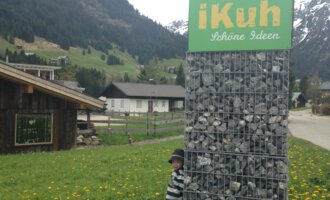 Ein Kind steht neben einem großen Schild mit der Aufschrift "iKuh" inmitten einer ländlichen Landschaft. Im Hintergrund sind Wiesen und Berge zu sehen. | © iKuh Schöne Ideen | Daniela Neumann