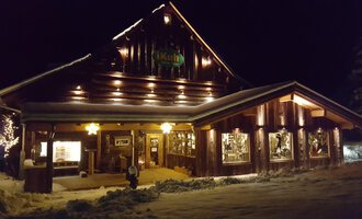 The shop iKuh Beautiful Ideas in a cozy wooden house at night, illuminated by warm light. It is surrounded by snow and radiates a cozy atmosphere.