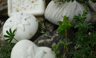 Handmade soaps in various shapes are lying on stones and grass. A small bag of dried flowers is also visible.