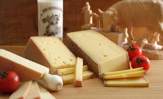 Ein Stück Käse liegt auf einem Holzbrett, umgeben von frischen Tomaten und Käse-Streifen. Im Hintergrund steht ein Glas Milch und eine Holzfigur eines Tieres. | © Innere Wiesalpe | Andrea Kainz zuckerschnecke.at