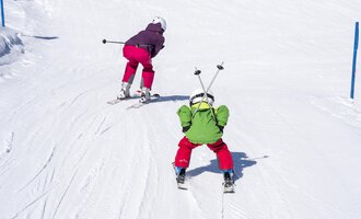 Zwei Kinder fahren Ski auf einer verschneiten Piste. Das eine Kind trägt eine grüne Jacke und das andere eine lila Jacke. | © Kleinwalsertal Tourismus | Dominik Berchtold