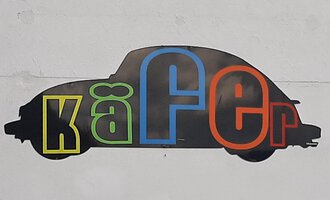 A stylized silhouette of a beetle car with colorful letters forming the word "Käfer." The background is simple and neutral. | © Kleinwalsertal Tourismus | V. Senn