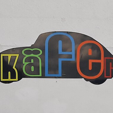 A stylized silhouette of a beetle car with colorful letters forming the word "Käfer." The background is simple and neutral. | © Kleinwalsertal Tourismus | V. Senn