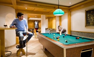 A cozy game room with a billiard table and a man watching relaxed. A woman is playing concentrated at the table. | © Aparthotel Kleinwalsertal Haller GmbH & Co KG
