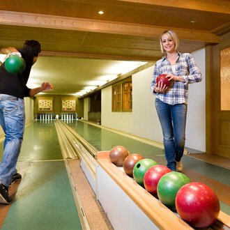 © Aparthotel Kleinwalsertal Haller GmbH & Co KG A cozy bowling alley with two people playing. One person throws a ball while the other stands on the lane holding a ball in hand. | © Aparthotel Kleinwalsertal Haller GmbH & Co KG