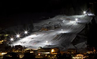 Eine beleuchtete Skipiste bei Nacht mit mehreren Skifahrern. Im Vordergrund sieht man gemütliche Hütten und eine ruhige Atmosphäre. | © Sport Kessler GmbH