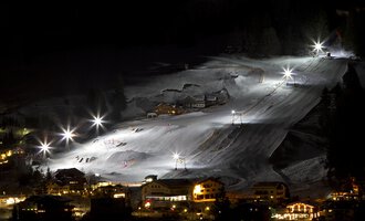 Eine verschneite Skilandschaft bei Nacht mit beleuchteten Pisten. Im Hintergrund sind kleine Hütten und ein klarer Himmel zu sehen. | © Sport Kessler GmbH