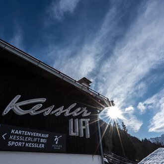 Ein gut sichtbares Schild mit dem Namen "Kessler" an einem Vordergrundgebäude. Im Hintergrund sind der Himmel und die Berge zu sehen. | © Sport Kessler GmbH