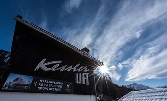 Ein gut sichtbares Schild mit dem Namen "Kessler" an einem Vordergrundgebäude. Im Hintergrund sind der Himmel und die Berge zu sehen. | © Sport Kessler GmbH
