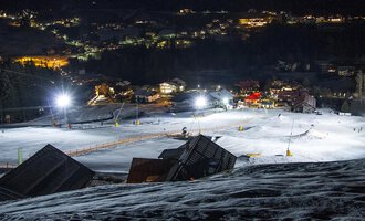 Eine beleuchtete Skipiste bei Nacht mit schneebedeckten Hängen. Im Hintergrund sind die Lichter eines Dorfes sichtbar. | © Sport Kessler GmbH