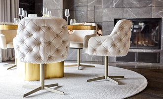 Ein eleganter Esstisch mit gepolsterten Stühlen in hellem Design. Im Hintergrund ist ein Kamin zu sehen, der eine gemütliche Atmosphäre schafft. | © Travel Charme Ifen Hotel