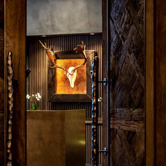 Ein stilvoller Eingangsbereich mit Holzwänden und einem dekorativen Geweih. Im Hintergrund ist ein Kunstwerk mit einem Tierkopf zu sehen. | © Travel Charme Ifen Hotel