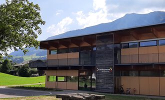 Ein modernes Gebäude mit großen Fenstern und Holzverkleidung. Im Vordergrund steht ein Steintisch auf einer grünen Wiese mit Bergen im Hintergrund. | © Kleinwalsertal Tourismus | Veronika Senn