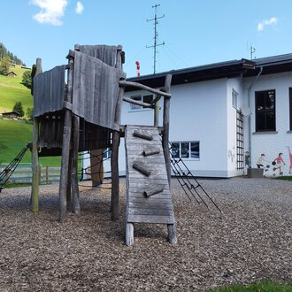 © Kleinwalsertal Tourismus | N. Lughammer A playground with a wooden climbing structure and a slide. In the background, you can see a house and green hills. | © Kleinwalsertal Tourismus | N. Lughammer