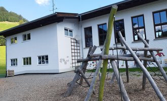 Ein modernes Gebäude mit großen Fenstern und einem Spielplatz aus Holz im Vordergrund. Der Spielplatz ist aus natürlichen Materialien und bietet verschiedene Spielmöglichkeiten. | © Kleinwalsertal Tourismus | N. Lughammer