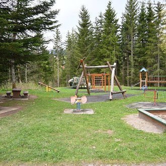 Ein Spielplatz in einem Wald mit mehreren Spielgeräten und einer Wiese. Im Hintergrund stehen hohe Bäume und eine Picknickbank. | © Gemeinde Mittelberg