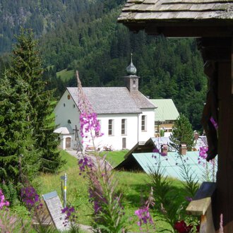 Eine malerische Landschaft mit einer Kirche und bunten Blumen im Vordergrund. Hohe Bäume und sanfte Hügel umgeben die Szene. | © Kleinwalsertal Tourismus | Rolf Köberle