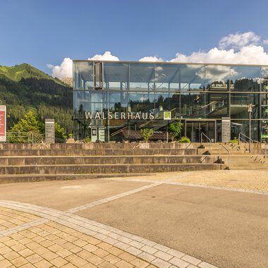 A modern building with a glass facade and the inscription "WALSERHAUS". In the background, green mountains and a blue sky are visible. | © Kleinwalsertal Tourismus | Steffen Berschin