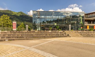 Ein modernes Gebäude mit Glasfassade und der Aufschrift "WALSERHAUS". Im Hintergrund sind grüne Berge und ein blauer Himmel sichtbar. | © Kleinwalsertal Tourismus | Steffen Berschin