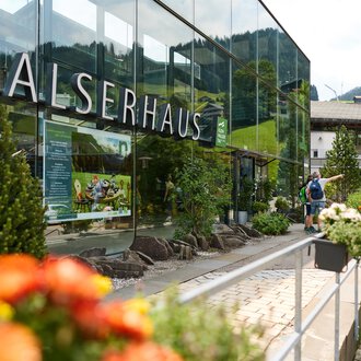 Ein modernes Gebäude mit Glasfassade und der Aufschrift "Walserhaus". Im Vordergrund blühen bunte Blumen, während im Hintergrund die Berge und traditionelle Gebäude zu sehen sind. | © Kleinwalsertal Tourismus | Oliver Farys