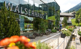Ein modernes Gebäude mit Glasfassade und der Aufschrift "Walserhaus". Im Vordergrund blühen bunte Blumen, während im Hintergrund die Berge und traditionelle Gebäude zu sehen sind. | © Kleinwalsertal Tourismus | Oliver Farys