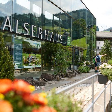 Ein modernes Gebäude mit Glasfassade und der Aufschrift "Walserhaus". Im Vordergrund blühen bunte Blumen, während im Hintergrund die Berge und traditionelle Gebäude zu sehen sind. | © Kleinwalsertal Tourismus | Oliver Farys