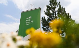 Ein Schild mit der Aufschrift "Klein Walsertal" steht im Vordergrund. Im Hintergrund sind grüne Bäume und bunte Blumen zu sehen. | © Kleinwalsertal Tourismus | Oliver Farys