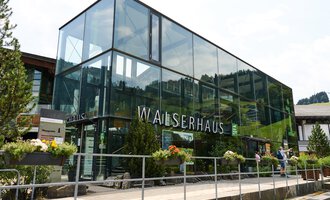A modern building with a glass front, surrounded by trees and flowers. In the foreground, there is a walkway where people are walking. | © Kleinwalsertal Tourismus | Oliver Farys