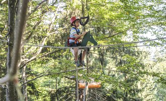 Ein Junge sitzt in einem Hochseilgarten auf einem Seil. Die Umgebung ist von Bäumen umgeben, und er trägt einen Helm und Sicherheitsausrüstung. | © OBERSTDORF · KLEINWALSERTAL BERGBAHNEN
