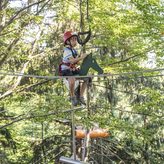 © OBERSTDORF · KLEINWALSERTAL BERGBAHNEN Ein Junge sitzt in einem Hochseilgarten auf einem Seil. Die Umgebung ist von Bäumen umgeben, und er trägt einen Helm und Sicherheitsausrüstung. | © OBERSTDORF · KLEINWALSERTAL BERGBAHNEN