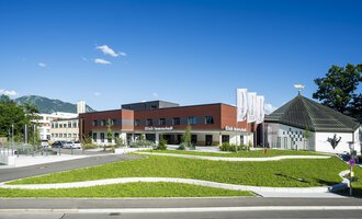 Ein modernes Gebäude mit einem geschwungenen Rasenbereich und einer klaren, blauen Himmel. Im Hintergrund sind Bäume und Berge sichtbar. | © Klinikverbund Allgäu