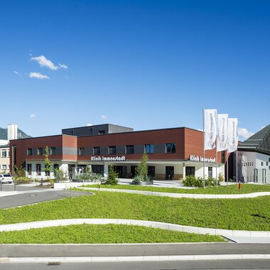 Ein modernes Gebäude mit einem geschwungenen Rasenbereich und einer klaren, blauen Himmel. Im Hintergrund sind Bäume und Berge sichtbar. | © Klinikverbund Allgäu