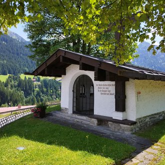 A small, white building with a gabled roof stands in a green landscape. In the background, gentle hills and trees can be seen. | © Kleinwalsertal Tourismus | Nicole Lughammer