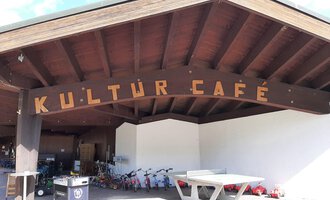 A wooden overhang with the inscription "KULTÜR CAFÉ" is visible. In the background, playground equipment and bicycles can be seen. | © Kleinwalsertal Tourismus | Veronika Senn