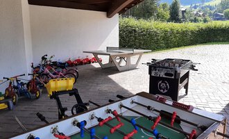 An outdoor area with a foosball table and various toys. In the background, playground equipment and a green landscape are visible. | © Kleinwalsertal Tourismus | Veronika Senn