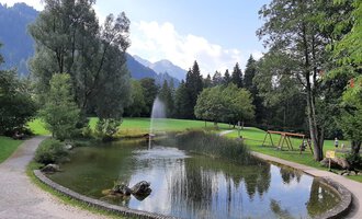 A quiet park with a pond and a fountain. In the background, trees and mountains can be seen. | © Kleinwalsertal Tourismus | Veronika Senn