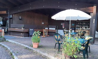 A cozy outdoor area with tables and chairs under a sunshade. In the background, wooden walls and shelves can be seen. | © Kleinwalsertal Tourismus | Veronika Senn