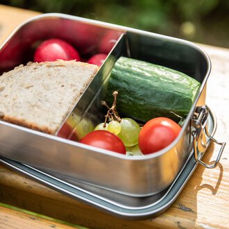 © Kleinwalsertal Tourismus | Bastian Morell A lunchbox with fresh bread, cucumber, cherry tomatoes, grapes, and radishes. It is on a wooden surface in a natural setting. | © Kleinwalsertal Tourismus | Bastian Morell