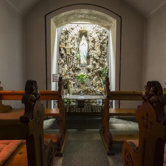 © Kleinwalsertal Tourismus | Oliver Farys Eine stille Kapelle mit Holzbänken und einer Statue in der Nische. Die Wände sind sanft beleuchtet und schaffen eine friedliche Atmosphäre. | © Kleinwalsertal Tourismus | Oliver Farys