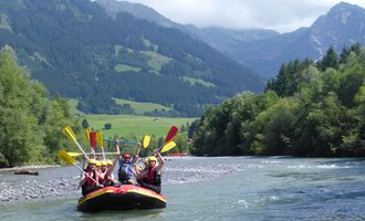 Eine Gruppe von Menschen paddelt im Schlauchboot auf einem Fluss. Die umliegende Landschaft ist grün und bergig, mit klarem Himmel im Hintergrund. | © MAP-Erlebnis