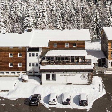 A snow-covered landscape with several buildings in chalet style. Surrounded by tall, snow-covered fir trees. | © Marburger Haus | Sweetchili