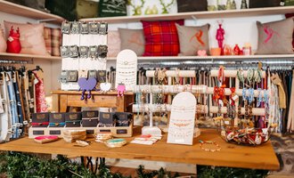A cozy shop with a variety of jewelry pieces and accessories on a wooden table. In the background, decorative pillows and clothing are visible. | © Herzstichle | Ninja Matt
