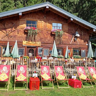 © Maxhütte | Viera Krafcik Ein traditionelles Holzhaus umgeben von Bäumen. Vor dem Haus stehen Liegestühle und Sonnenschirme in einer freundlichen Atmosphäre. | © Maxhütte | Viera Krafcik